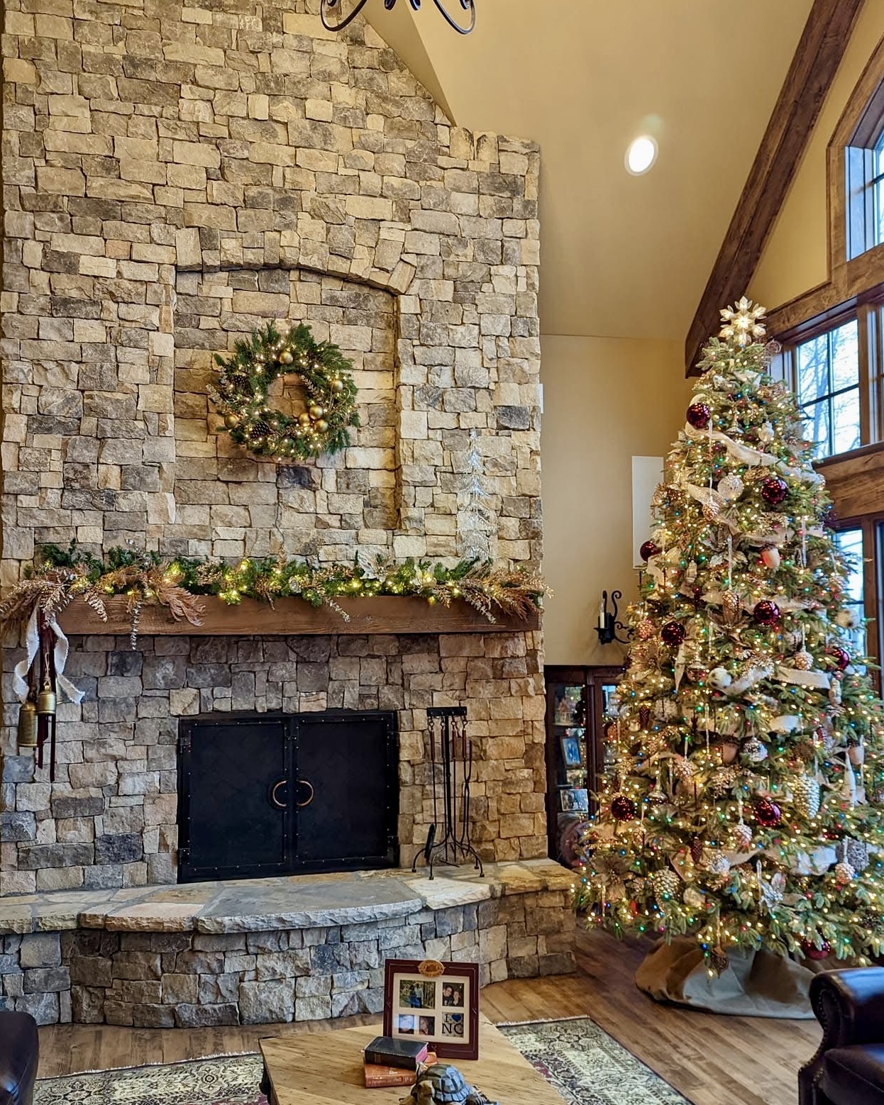A stone hearth with a golden wreath, lit garland, and a tree in burgundy and gold